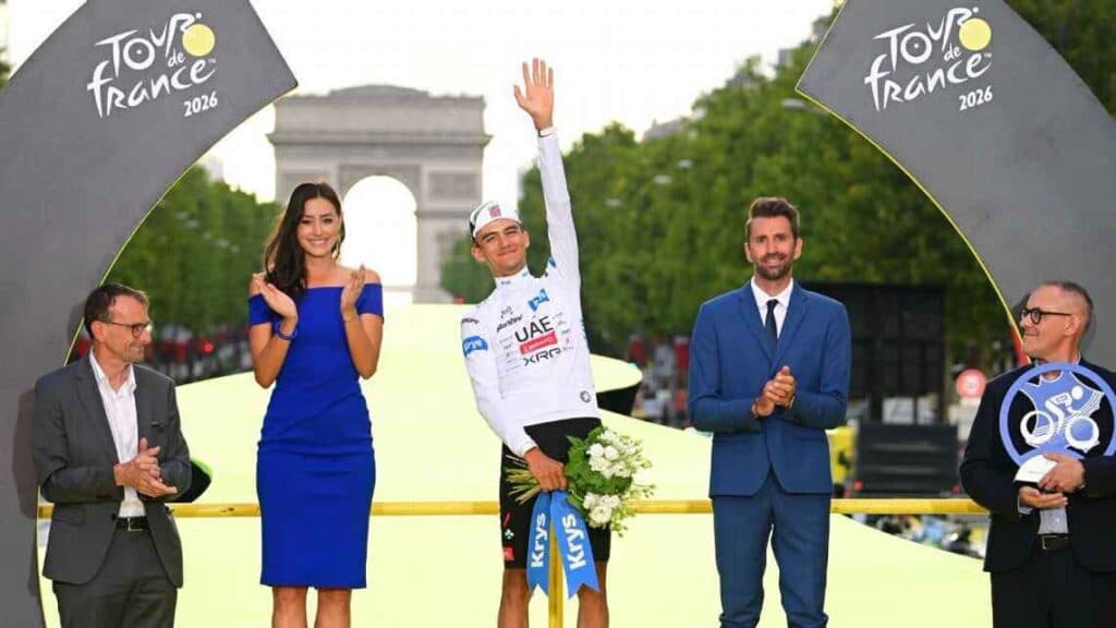 Isaac del Toro, en el podio del Tour de Francia 2026, saluda mientras sostiene flores, rodeado de autoridades y el Arco del Triunfo al fondo.